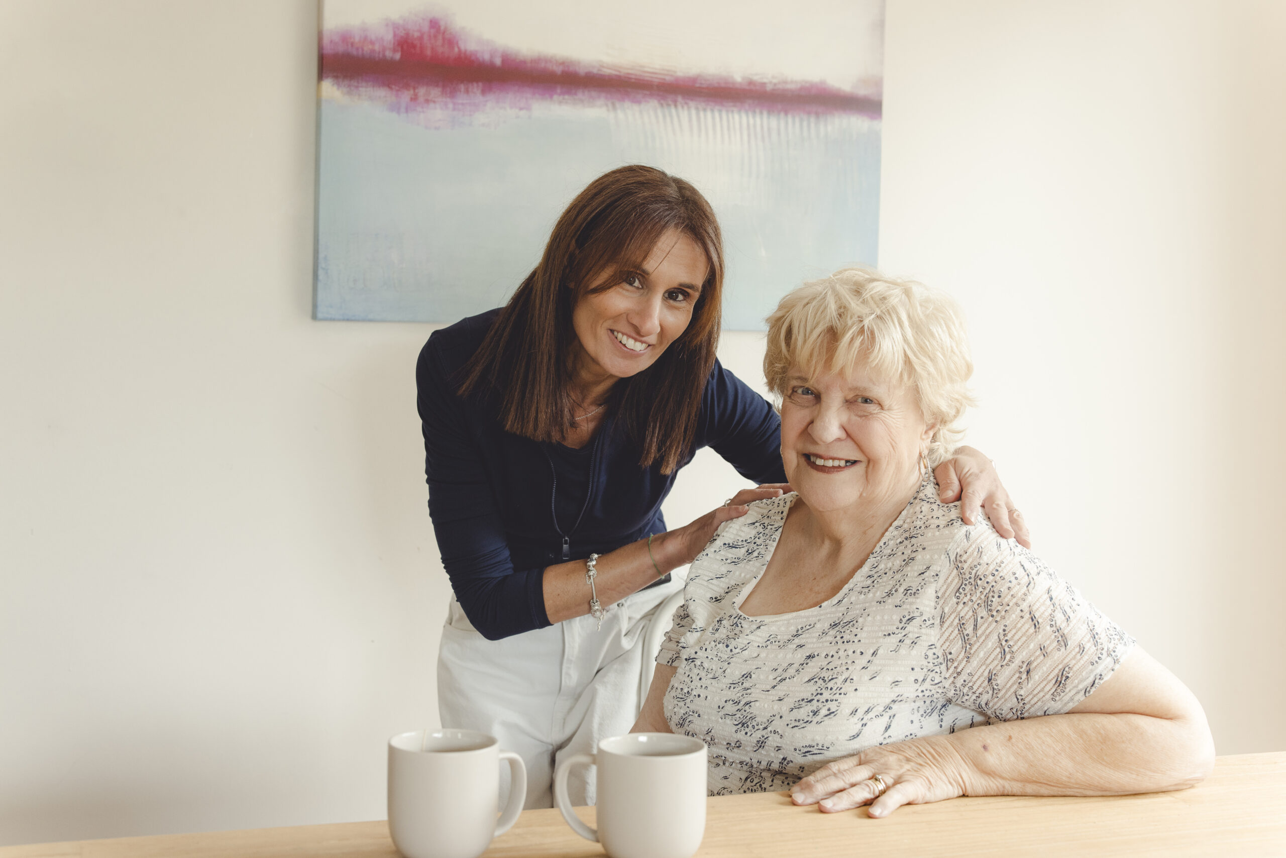 A young woman standing beside a senior woman who is seated at a table