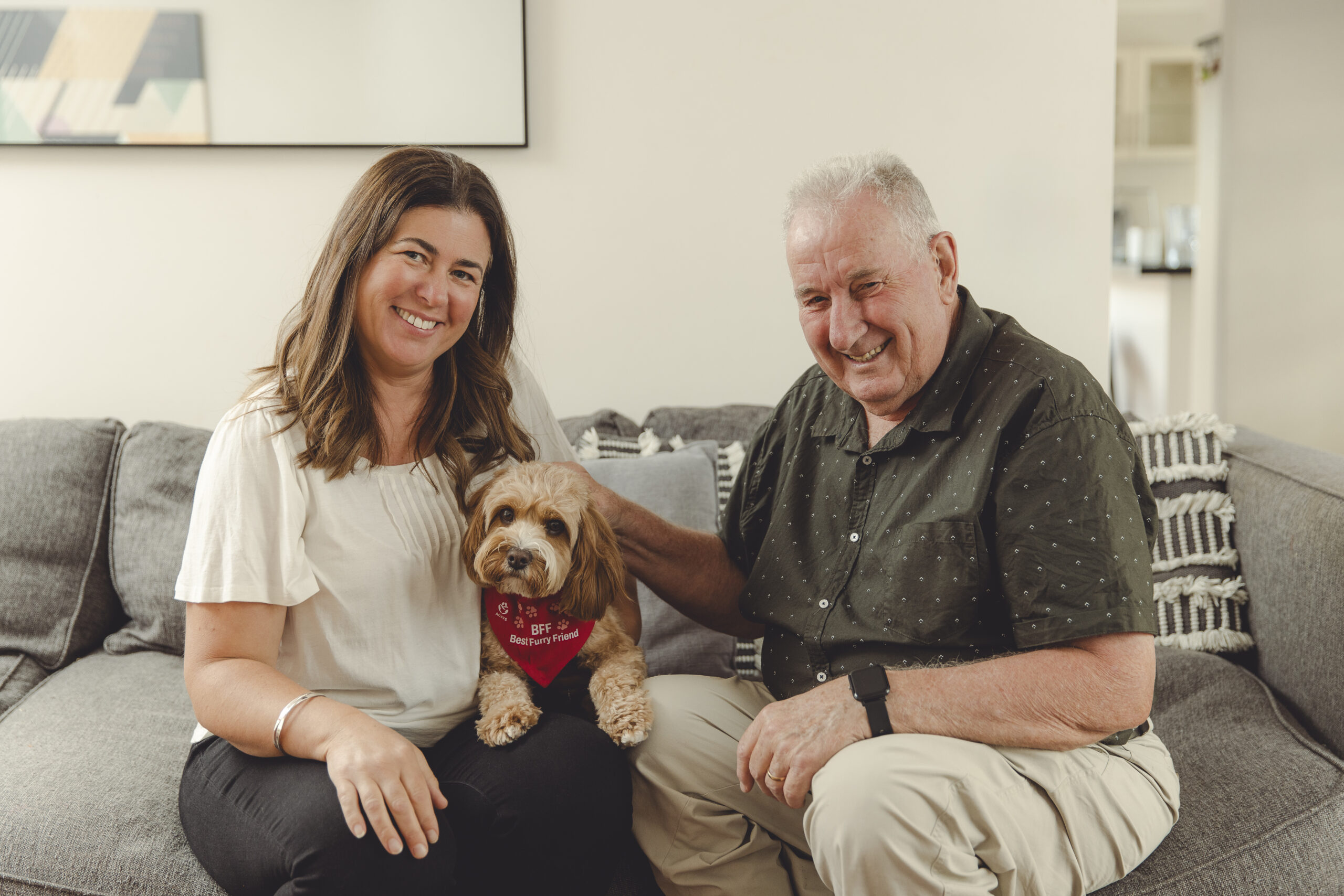 A woman with a small dog is seated on the couch beside a senior man